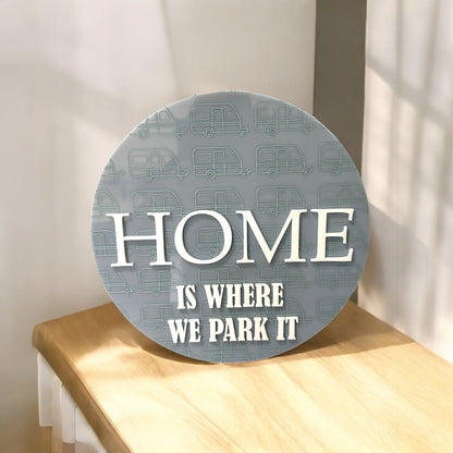 Round decorative sign with 'HOME IS WHERE WE PARK IT' text on a wooden surface.
