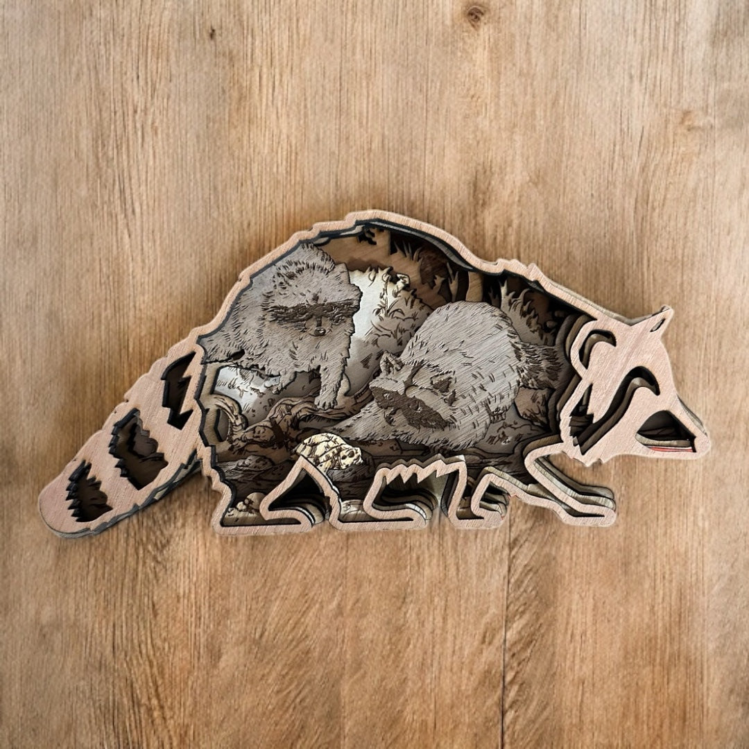 Layered wooden raccon with two raccoons inside in on a forest floor.