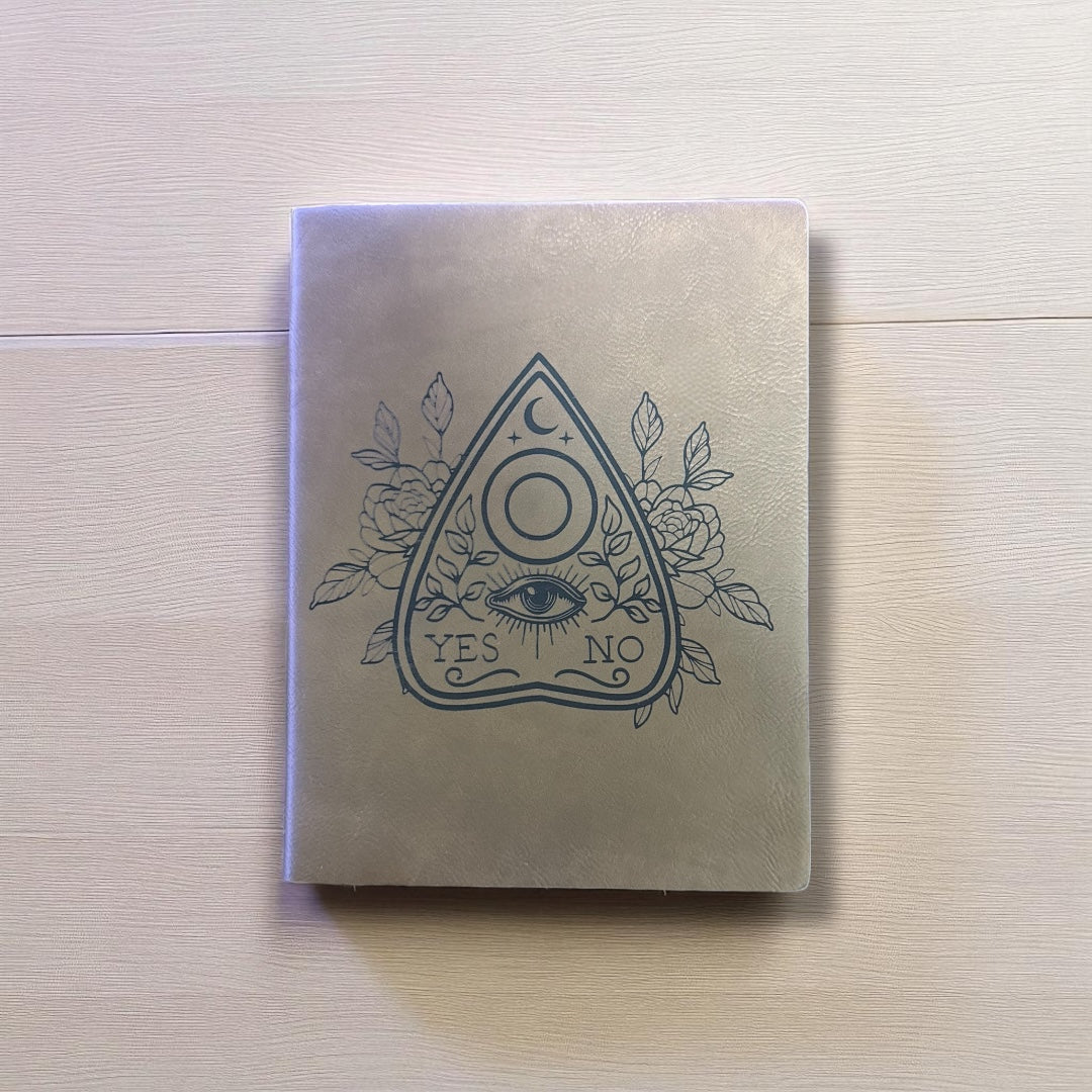 Notebook with a mystical design on the cover, featuring an eye, moon, and roses, on a light wooden surface.