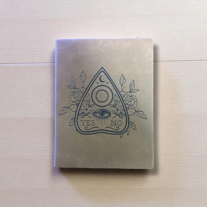 Notebook with a mystical design on the cover, featuring an eye, moon, and roses, on a light wooden surface.