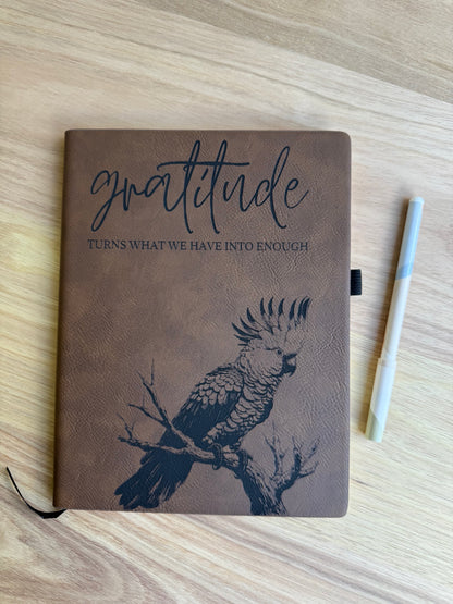 Brown notebook with a parrot illustration and 'gratitude' quote on a wooden surface.